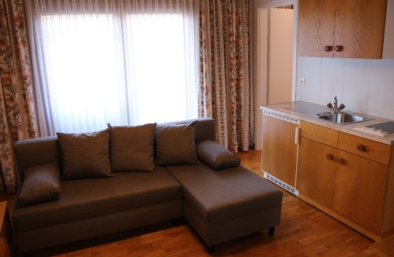 Living room with gray sofa in front of window with floral curtains and adjacent wooden kitchenette