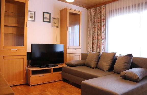 Living room with gray corner sofa on right, wooden cabinets and TV on left, window with curtains in background