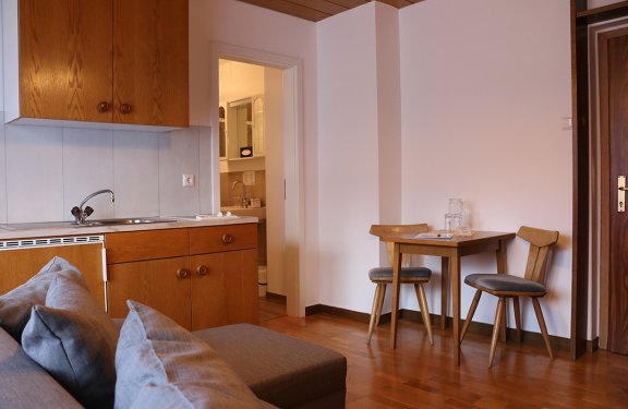 Small kitchen with wooden cabinets on left, gray sofa in foreground, table with two chairs on right