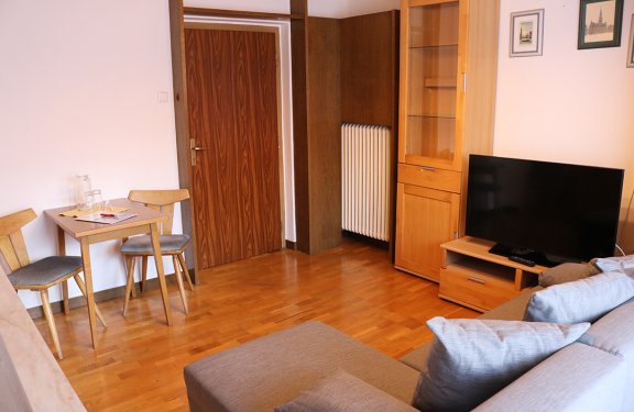 Living room with gray sofa on right, wooden table with two chairs on left, and TV on wooden cabinet